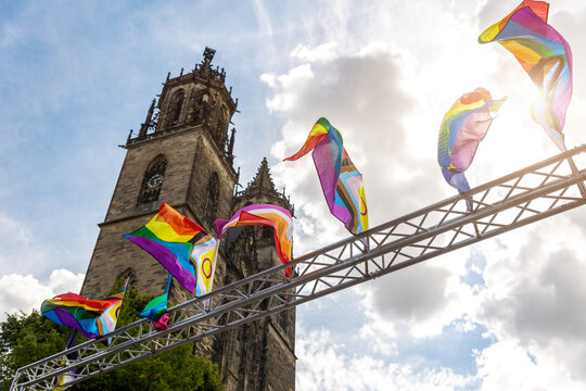 Rainbow and inclusive pride flags wave on metal truss beside historic cathedral Dom towers Magdeburg. Colorful banners symbolize LGBTQ visibility, diversity, equality, solidarity and celebration