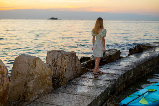 Fashionable Girl Standing on Pier at Sunset. Fashionable girl in a floral dress standing on a stone pier by the sea at sunset, with coastal rocks and a paddleboard nearby.