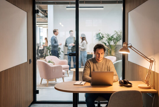 Man works on laptop in office while colleagues talk in background