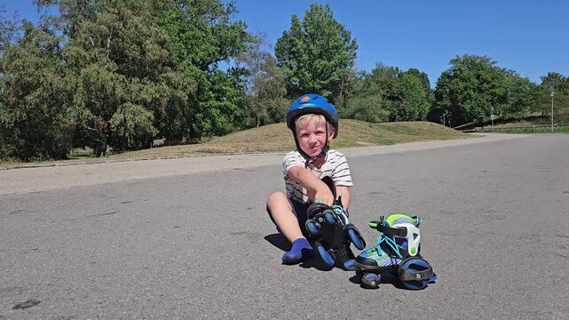 A cheerful toddler in a striped tee and navy shorts sits cross‑legged on sunlit asphalt, wearing a bright blue safety helmet; a teal‑and‑green rollerblade rests beside him, while lush trees and a vivi