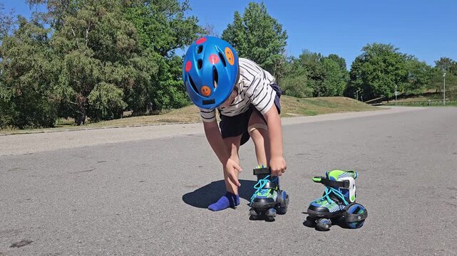 A young boy in a striped tee and navy shorts crouches on sunlit asphalt, adjusting his blue safety helmet; his teal-and-green rollerblades rest nearby, while lush trees and a vivid blue sky frame the 