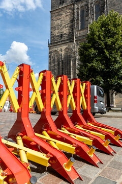 Portable red and yellow security barriers stand on cobblestone square beside historic Gothic church. Heavy mobile roadblocks provide temporary vehicle access control during event in Magdeburg