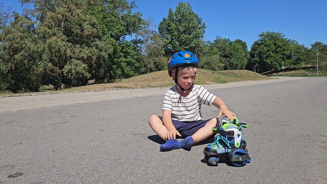 A calm boy in a striped tee and a bright blue safety helmet sits on asphalt, looking at the camera: his face shows joy, calmness, or mild fatigue; rollerblades are nearby, framed by green trees and a 