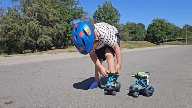 A smiling boy in a striped tee and a bright blue safety helmet poses on sunlit asphalt, interacting with his rollerblades, surrounded by lush trees and a bright blue sky. The scene shows joyful posing