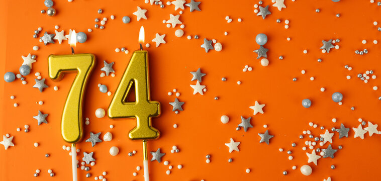 Birthday or anniversary celebration concept. Candles in the shape of the number seventy-four on an orange background among decorative ornaments. 74th anniversary. Banner