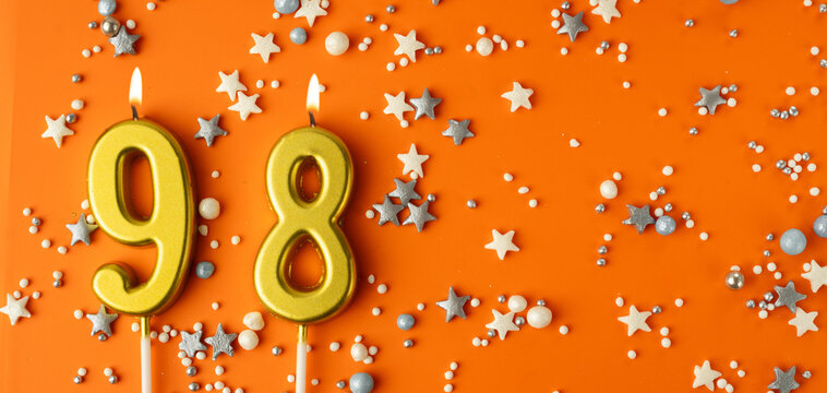 Birthday or anniversary celebration concept. Candles in the shape of the number ninety-eight on an orange background among decorative ornaments. 98th anniversary. Banner