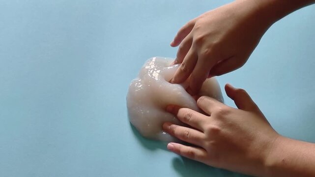 Child's hands playing with clear glitter slime for asmr and sensory stimulation, showing a satisfying tactile texture experience on a solid light blue background
