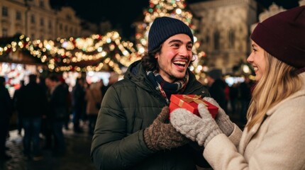 Fototapeta premium Woman giving Christmas gift box to happy man at night winter market. Holiday present exchange with illuminated tree and festive lights. Christmas and New Year celebration concept