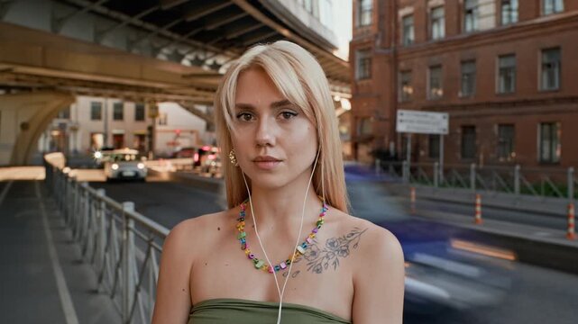 Portrait of young adult Caucasian woman wearing earphones, standing still on city street with time lapse traffic in background. Footage is perfect for music streaming or urban lifestyle commercials