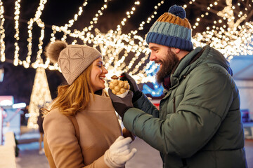 Obraz premium Couple feeding each other with sweet snacks while enjoying winter holiday season outdoors in the city