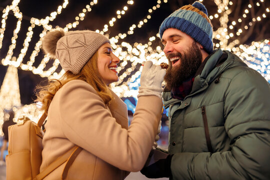 Couple feeding each other with sweet snacks while enjoying winter holiday season outdoors in the city