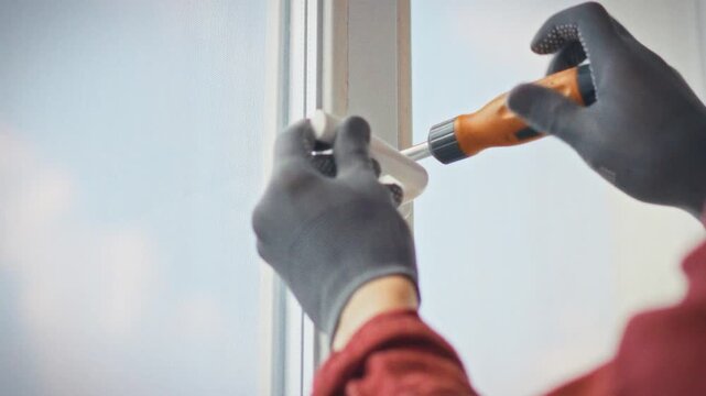 A close-up of an orange screwdriver in gloved hands carefully removing a handle from a plastic window frame at home. Concept of precise and professional window handle repair and maintenance