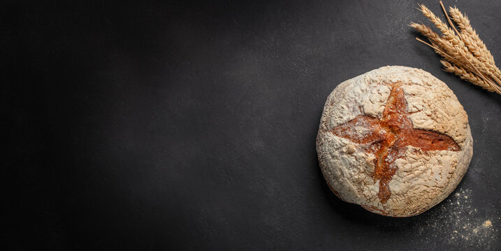 Round artisanal bread loaf with a cross pattern on a dark surface, accompanied by wheat stalks, showcasing rustic baking and natural ingredients. copy space