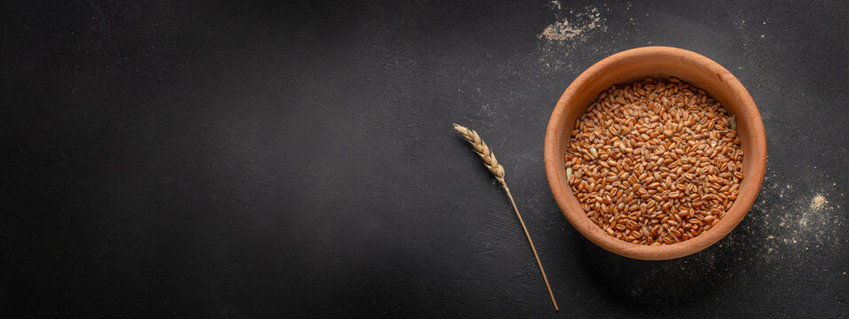Bowl of whole wheat grains on a dark surface with a single wheat stalk beside it, showcasing natural food ingredients in a rustic setting. copy space