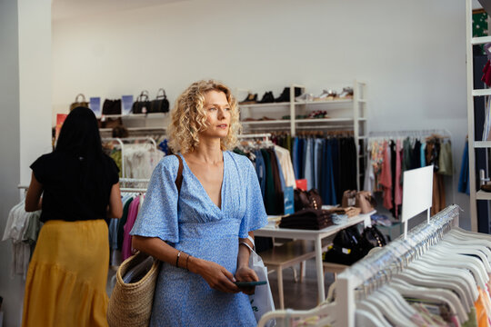 Blonde Woman Shopping in Fashion Boutique Wearing Blue Summer Dress and Carrying Straw Bag