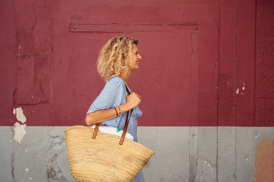 Smiling Blonde Woman Carrying Large Straw Shopping Bag Along Colorful Mediterranean Street