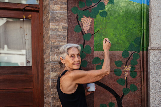 Senior Female Artist Painting Decorative Grape Vine Mural on Exterior Wall in Spanish Town