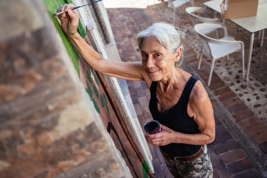 Smiling Senior Artist Looking at Camera While Painting Outdoor Wall Mural with Fine Brush