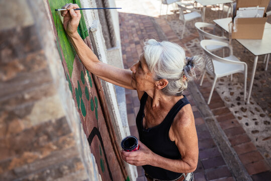 Senior Female Artist Painting Colorful Outdoor Wall Mural from Above During Creative Street Project