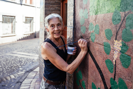Smiling Senior Woman Painting Decorative Grape Vine Mural on Traditional Street Building Facade