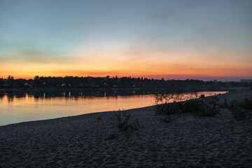 Sunset over a calm river with orange and blue sky reflected on the water, sandy shoreline in foreground and distant trees. Natural evening landscape with copy space for travel themes © Photolove