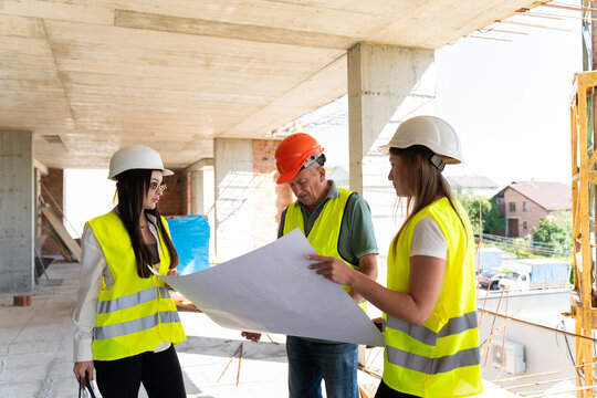Project managers and builders study large floor plan. Female engineers hold clipboard discussing work process with builder in safety hat