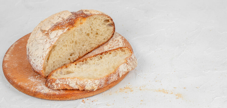 Freshly baked round sourdough bread loaf sliced on a wooden cutting board with flour dust and a light gray background for culinary and food photography. copy space