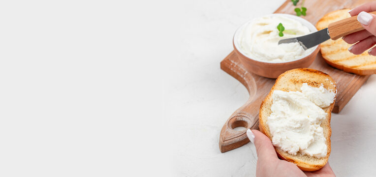 Hand spreading creamy cheese on toasted bread slice with knife, bowl of cheese and fresh herbs on wooden cutting board in bright kitchen setting. copy space.