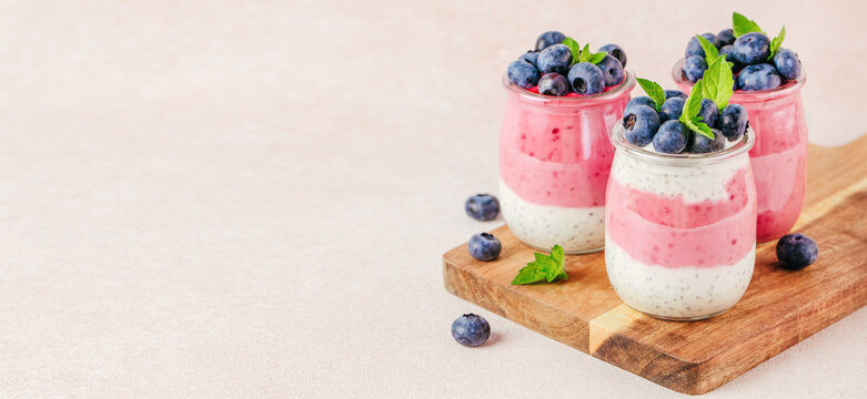 Three layered yogurt desserts with pink and white stripes topped with fresh blueberries and mint leaves on a wooden cutting board against a light background. copy space