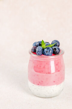 Layered dessert in glass jar featuring pink yogurt, chia seeds, topped with fresh blueberries and mint leaves against a soft pastel background. copy space