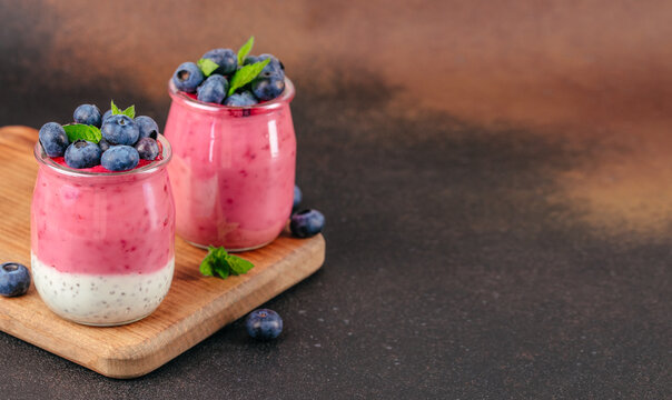 Two glass jars filled with layered yogurt dessert topped with fresh blueberries and mint leaves on a wooden cutting board against a dark background. copy space