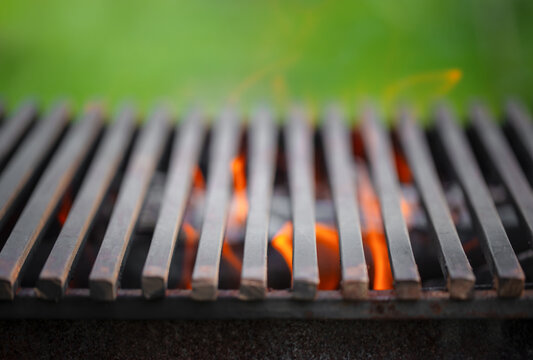 Close-up view of a metal grill with flames visible beneath the slats, set against a blurred green background, highlighting outdoor cooking equipment