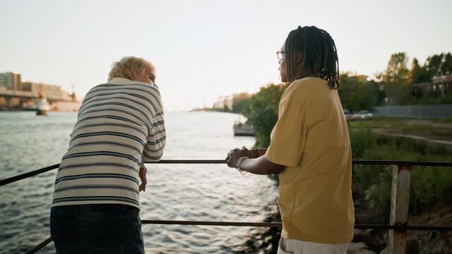 Diverse young adults, one Caucasian and one Black, leaning on railing by river at sunset and talking. Clip is perfect for multicultural friendship and outdoor lifestyle commercial projects