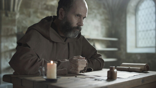 Bearded medieval scribe looking away thoughtfully while holding a rosary at a rustic wooden table inside a scriptorium