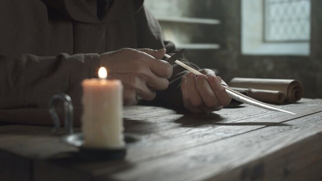 Hands of a medieval monk cutting a feather quill pen with a knife at a wooden table next to a burning candle inside a scriptorium