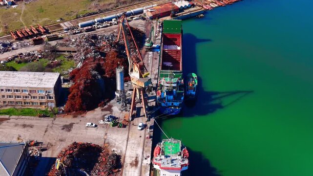 Aerial view of scrapyard crane loading cargo ship, port. Overhead shot shows rusty metal pile, dockside crane and green harbor water.
