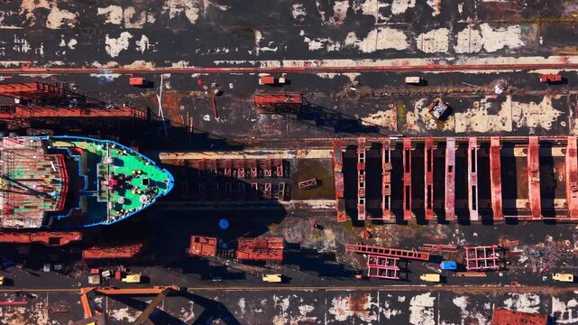 Overhead view of ship bow in shipyard dry dock. Green deck, rusted rail tracks and scattered steel beams.