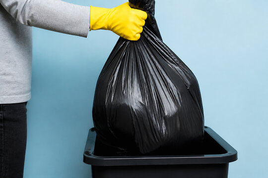 A person in a grey shirt and yellow gloves is holding a black trash bag over a black bin against a light blue background.