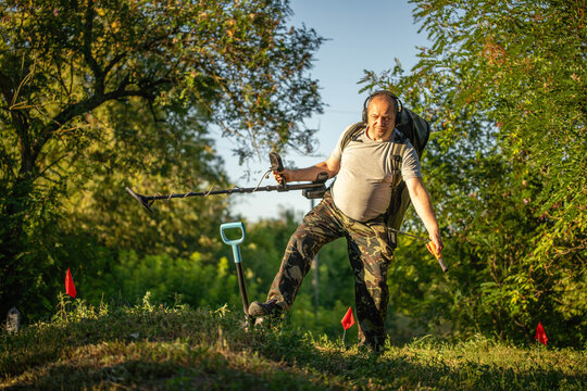 A man searches for metal objects in the forest with a metal detector. He stands on a small hill and holds a shovel in one hand. The sun is setting in the background
