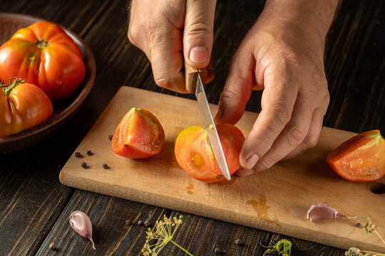 A chef prepares to slice a ripe tomato on a wooden cutting board in a kitchen. Fresh garlic and herbs are placed around the cutting board. The scene shows food preparation