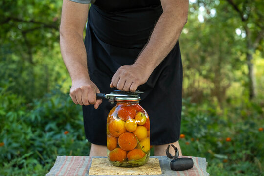 A cook is using a sealing machine to close a large jar filled with tomatoes. This is happening in a garden with green plants and trees around