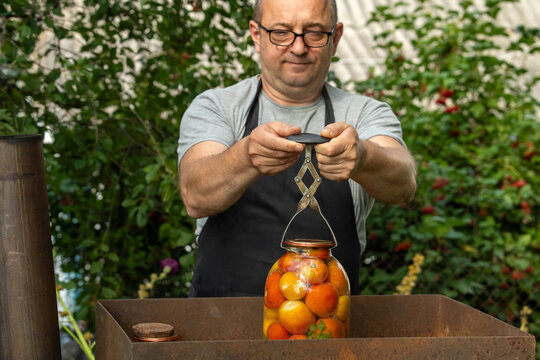 A man stands in his garden weighing a jar filled with tomatoes. The scene shows greenery and sunlight. He uses a scale to measure the jar's weight
