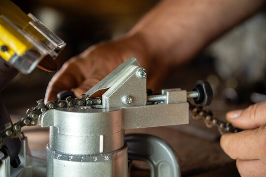 A master is using a chain sharpener in a workshop. The tool is being carefully adjusted to sharpen the chain. Light comes through the workspace, showing the details of the task