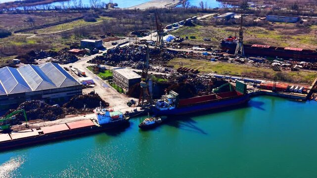 Aerial view of port scrapyard with cargo ship, Bulgaria. Metal scrap piles, warehouse, dockside crane and green harbor water.
