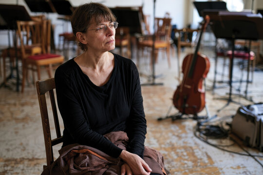 A pensive female musician sits in a performance space, surrounded by chairs and musical instruments, embodying the emotions tied to the art of music and performance.