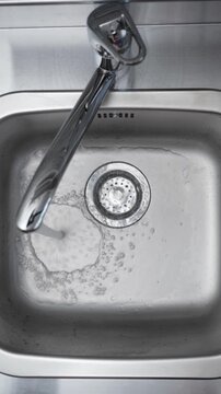 Hand Turning On Faucet to Fill Kitchen Sink. Top view of a person opening a chrome tap as running water begins filling a stainless steel sink.