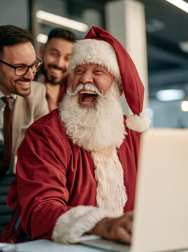 Santa Claus laughing loudly while sitting at desk with laptop surrounded by smiling office colleagues