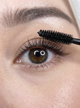 Close up view of a young woman applying mascara to her upper eyelashes with a black brush applicator, emphasizing eye makeup and beauty routine.
