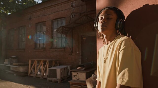 African American young adult wearing headphones leaning against wall outdoors, eyes closed, listening to music in slow motion. Footage suits youth lifestyle and audio technology marketing campaigns