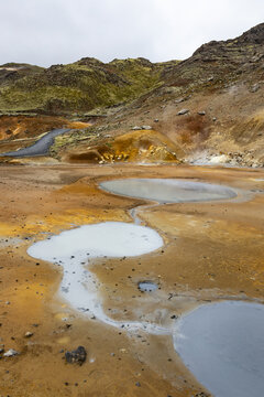 View of Seltun Geothermal Area with bubbling mud pools, steaming vents, and colorful mineral deposits against a backdrop of rugged volcanic hills in Grindavik, Hafnarfjardarkaupstad, Iceland.